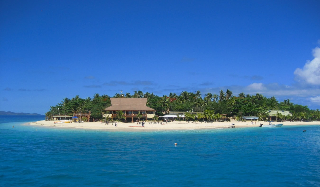 A white sand beach on an island surrounded by deep blue water and covered in palm trees on a sunny day