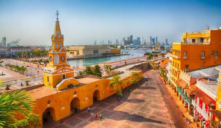 A view over an expansive plaza surrounded by bright orange historic buildings with the harbor and modern skyscrapers in the background in Cartagena, Colombia