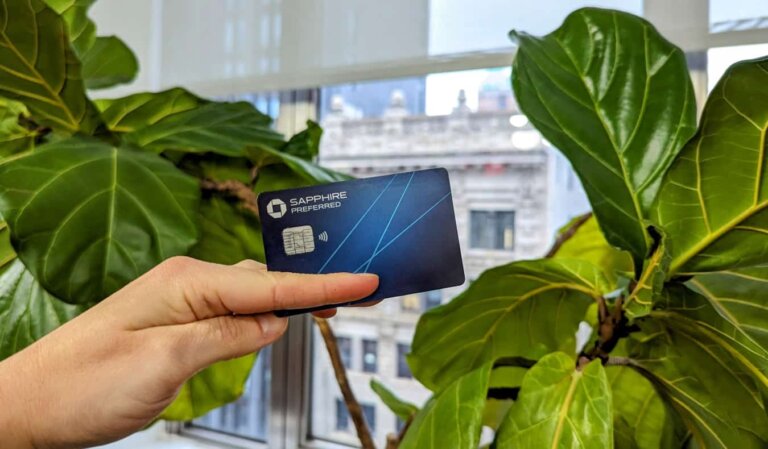 The Chase Sapphire Preferred Card held up before a leafy plant with buildings seen through the window in the background