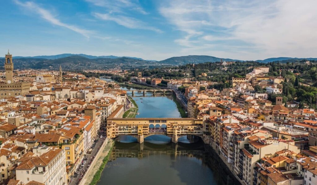 Aerial view of the city of Florence with the Arno River running through it, with Ponte Vecchio in the foreground and mountains in the background