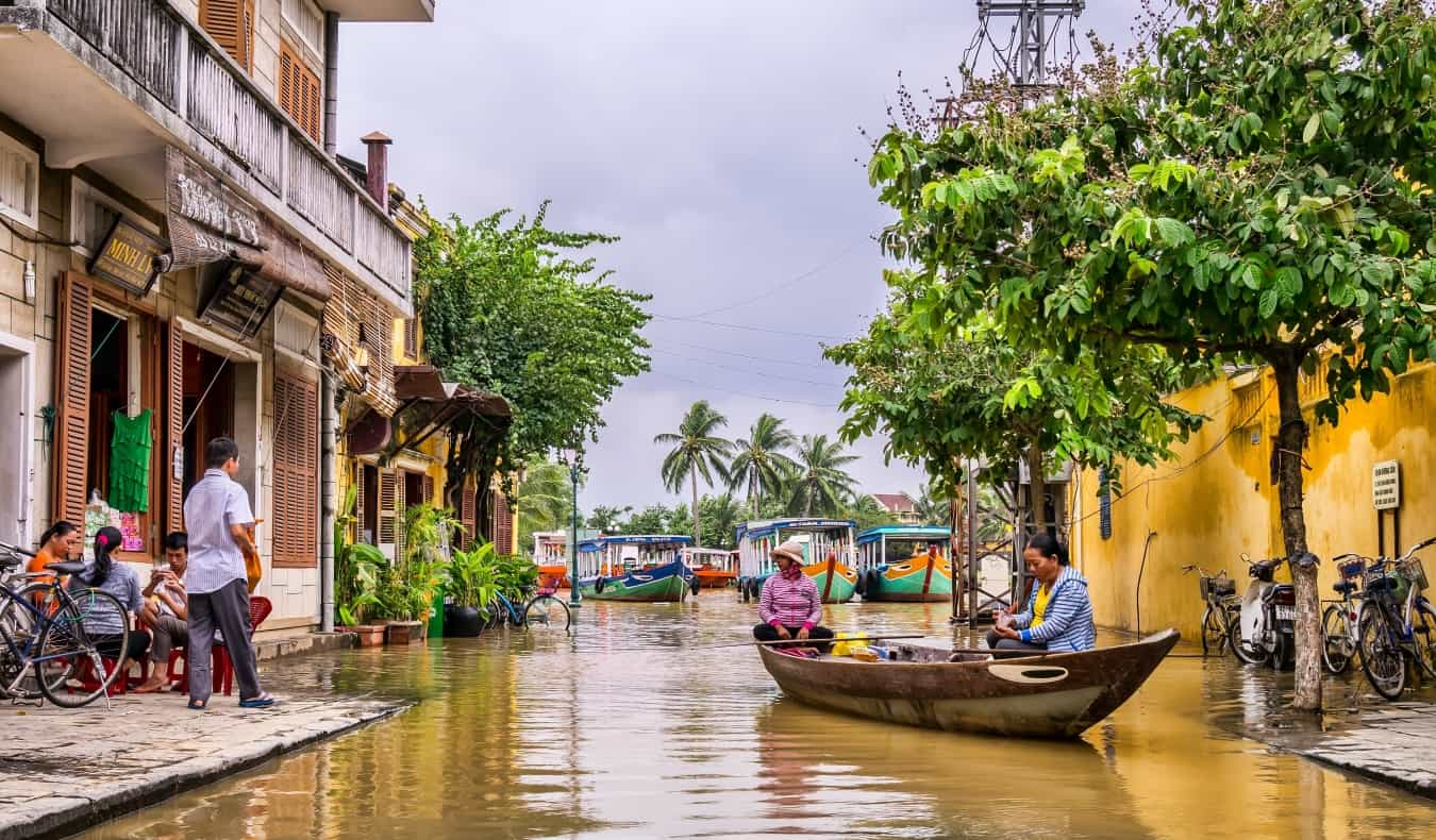 Women sitting in a boat with people on the sidewalk of buildings nearby in Hoi An, Vietnam