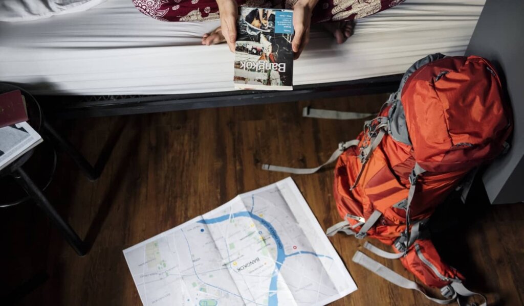 A person sits on a hostel bunk, holding a guidebook to Bangkok in their hands, with their backpack nearby on the ground