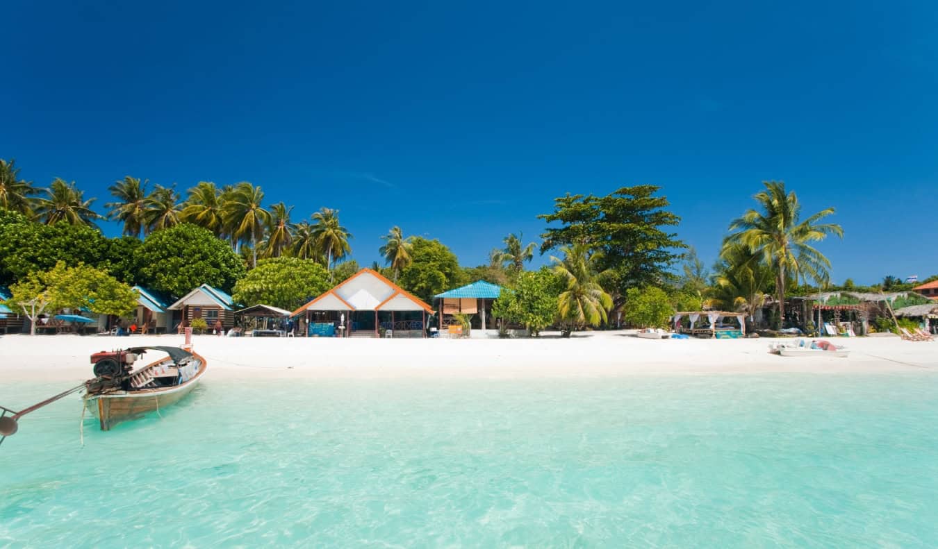 Bungalows along a white sand beach with a boat floating off to the side on the island of Ko Lipe in Thailand