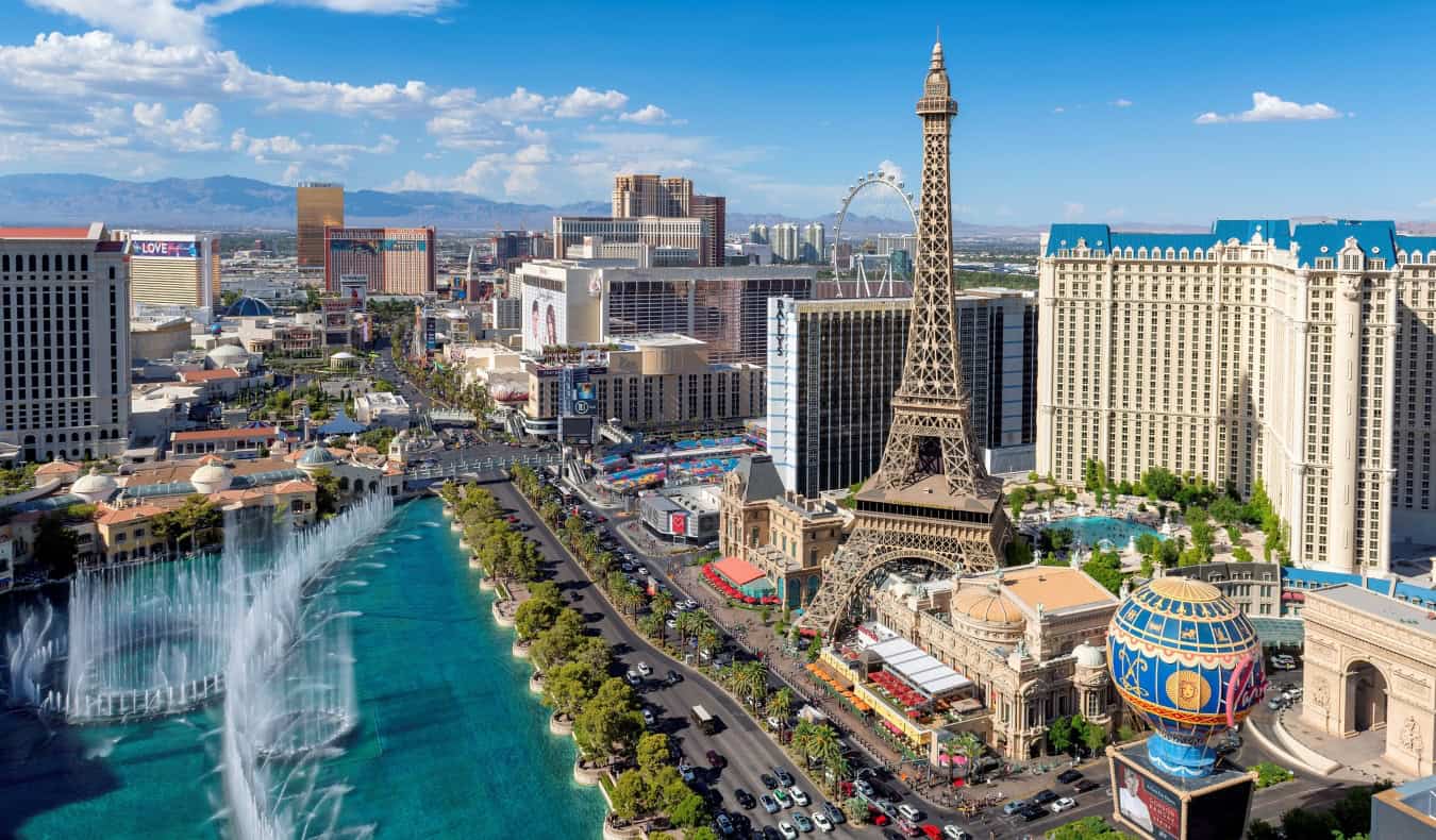 An aerial view of the Las Vegas strip, with extravagant fountains, the Eiffel Tower replica, and many other large hotels, with the mountains in the background