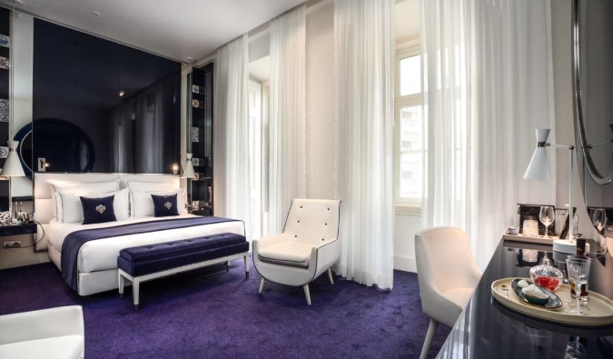 A luxurious suite decked out in deep blue rugs, a sleek desk, white armchair, large bed, and French doors covered in sheer curtains at Portugal Boutique Hotel in Lisbon, Portugal