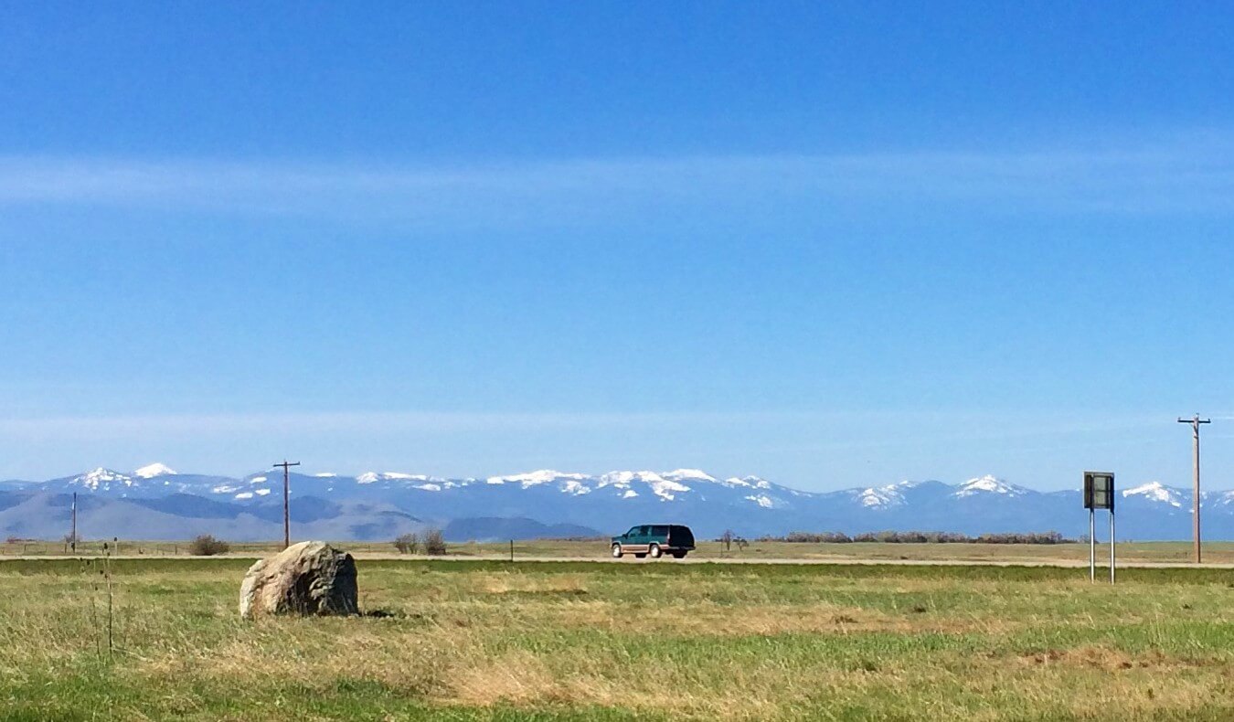 A car drives down an open road in front of fields with mountains in the background in Montana, USA