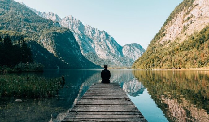 man sat on the end of a pontoon wearing black surrounded by a lake and mountains