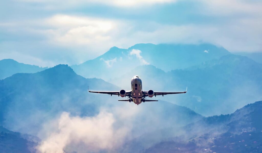 A lone commercial jet flying through a bright blue sky with mountains in the distance