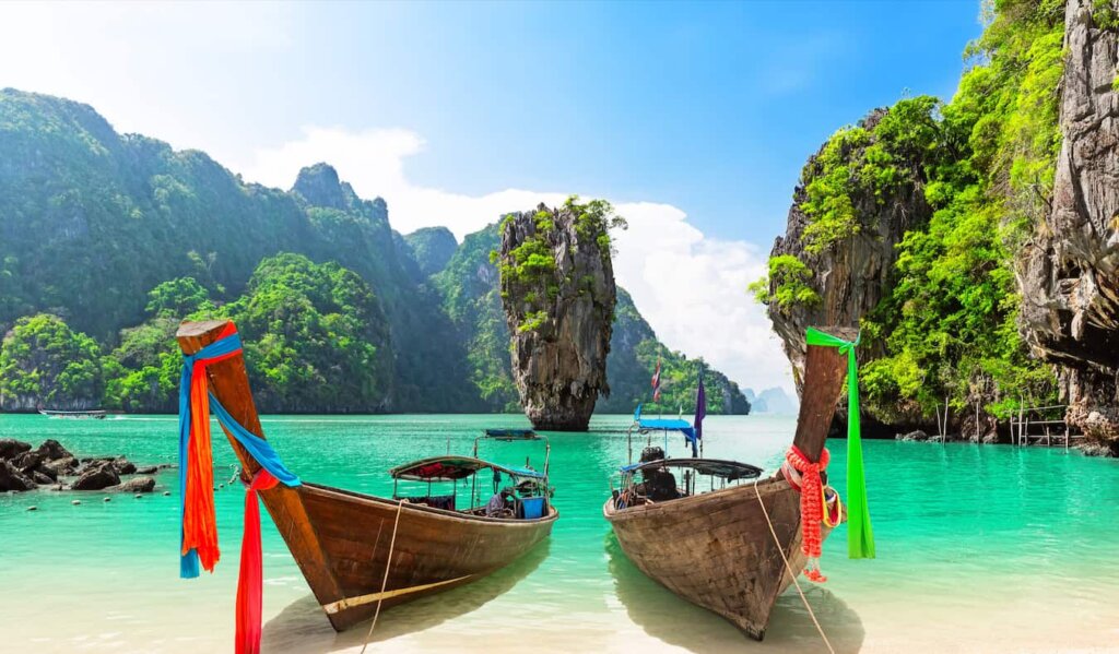 Red longtail boats pulled up to a picturesque beach with cliffs in the background, in Thailand